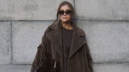 Woman standing against grey wall, wearing brown sweater and brown teddy fall coat, black glasses, and gold hoop earings.