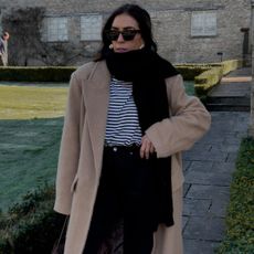 Influencer wears camel coat, striped shirt, black jeans, black scarf, and sunglasses.