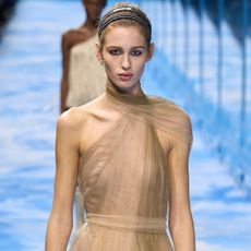 A woman wears a double black headband, and a gold sheer dress at the Dior S/S 25 show.