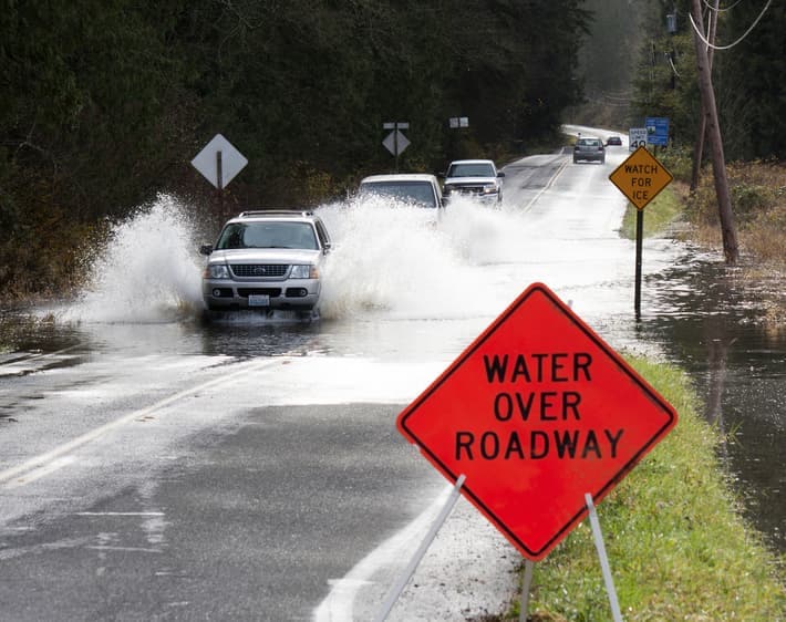 Line of Cars on Two-Way Road Driving Through Deep Puddle with Bright Orange "Water Over Roadway" Sign at Forefront of Image.