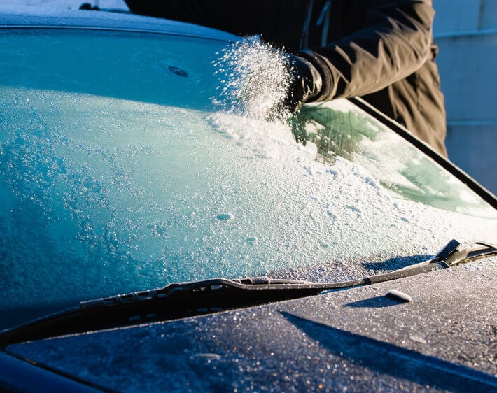 Man wearing gloves scraping windshield frost off his windshield in the morning sun