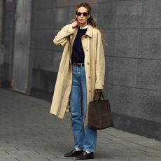 female fashion influencer Anouk Yve poses wearing oval sunglasses, a collared trench coat, a black sweater layered over a white tee, brown suede bag, belted jeans, white socks, and ruched loafers